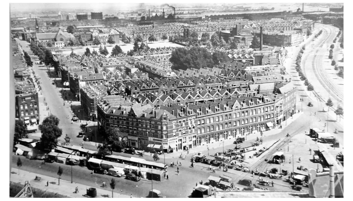 Zwart-witfoto van de Afrikaanderwijk in 1950, met rijen woonhuizen, een winkelstraat met marktkramen en een brede weg.