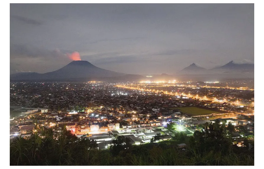 Nachtfoto van de stad Goma met op de achtergrond de vulkaan Nyiragongo, waarvan de top rood oplicht.