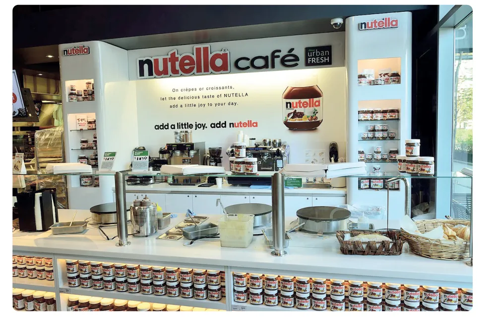 Foto van een Nutella-café met een toonbank, crêpeplaten en rijen potten Nutella in de vitrine.