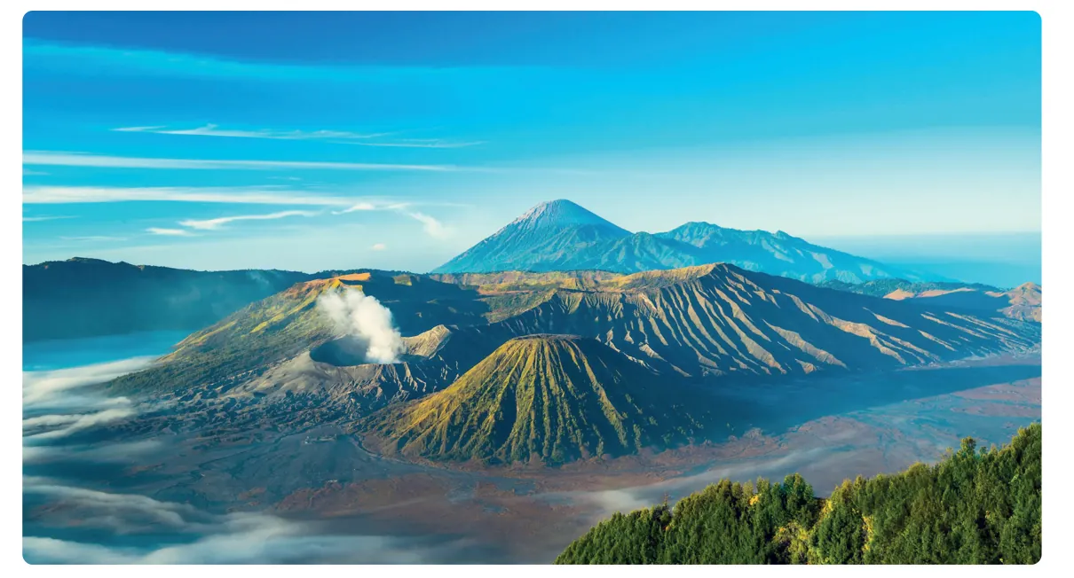 Foto van een caldera met een rokende krater en een kegelvormige vulkaan op een vlakte, met op de achtergrond een hoge, spitse vulkaan.