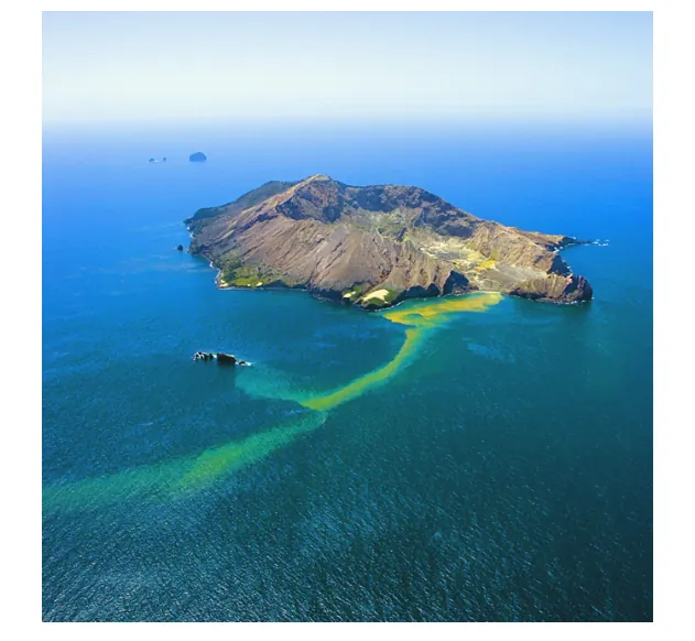 Luchtfoto van White Island: een kaal, vulkanisch eiland in zee, met een gele slierte water langs de kust.