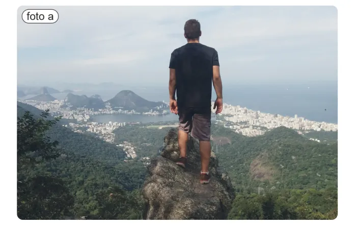 Een man staat op een rots boven een bebost berglandschap en kijkt uit over een kuststad aan een baai met bergen.