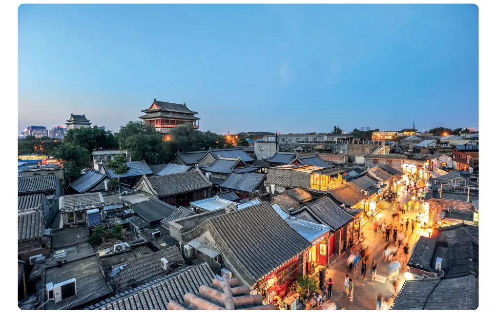 Avondfoto van een hutong in Beijing met lage daken van grijze tegels, een hoge poort op de achtergrond en verlichte winkels.