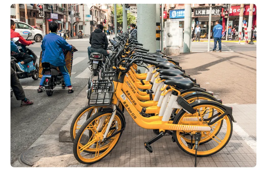 Rij oranje deelfietsen geparkeerd op de stoep langs een drukke straat in Shanghai, met voetgangers en scooters.