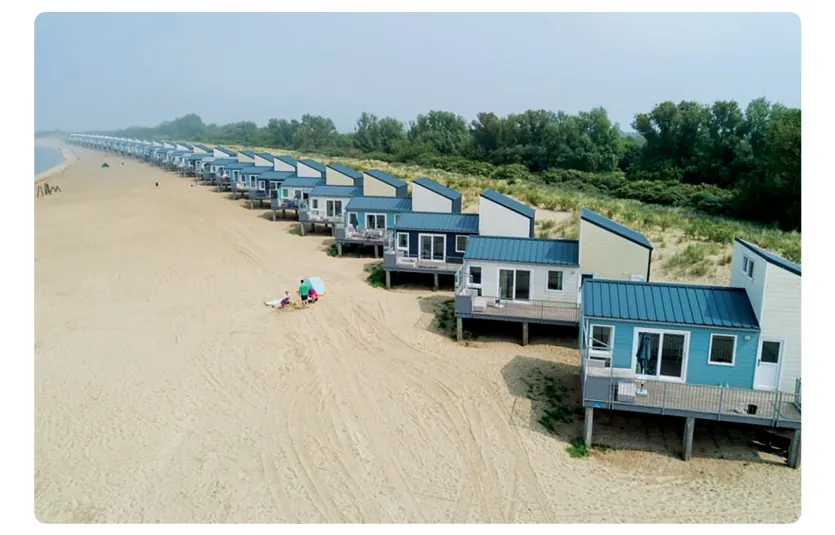 Foto van een lange rij strandhuisjes op palen op het strand, direct voor de begroeide duinen.