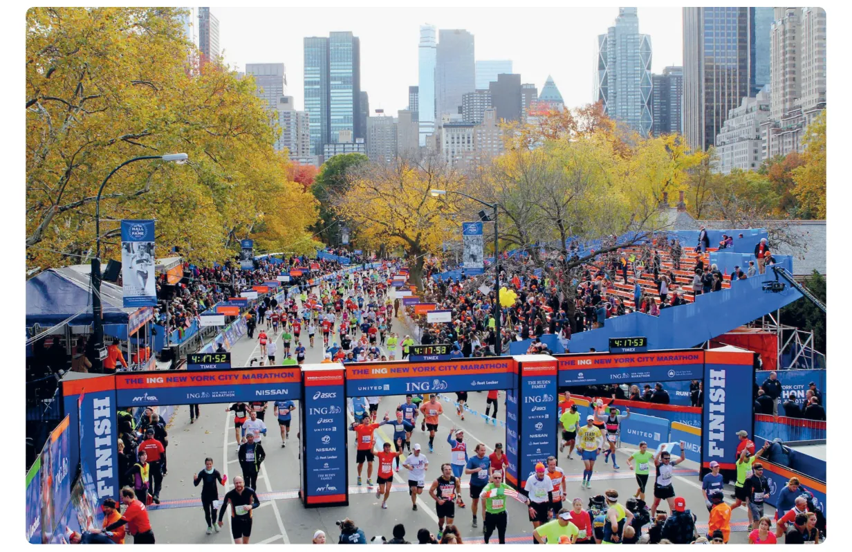 Foto van lopers die onder de finishbogen van de New York City Marathon door komen, in een park met herfstbomen en op de achtergrond de wolkenkrabbers van Manhattan.