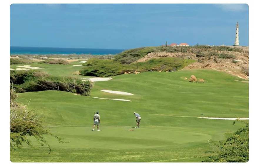 Foto van een golfbaan met groen gras, zandbunkers en droge struiken, met de zee op de achtergrond en een vuurtoren.