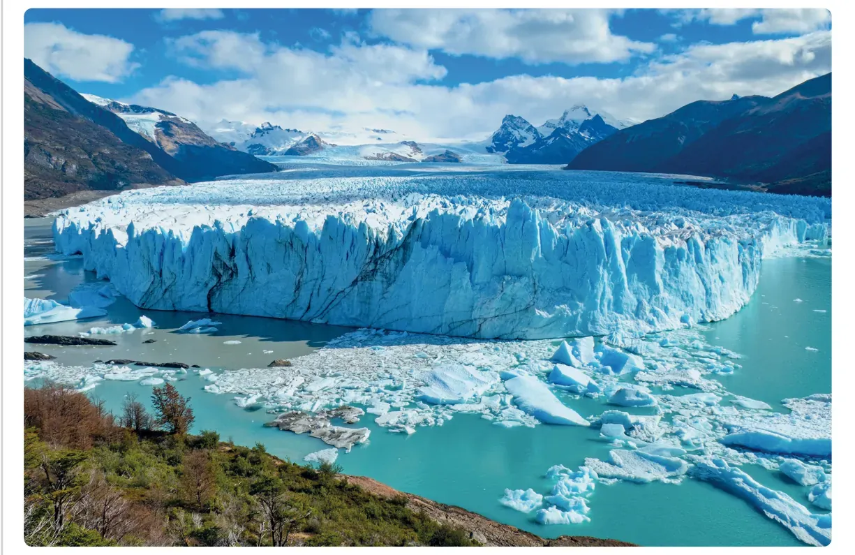 Foto van de Perito Moreno-gletsjer: een brede ijsmuur die uitmondt in turquoise water, met ijsschotsen en een begroeide oever op de voorgrond.