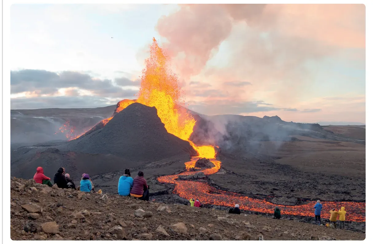 Foto van een lavafontein en een lavastroom op IJsland, met toeristen die op enige afstand toekijken.