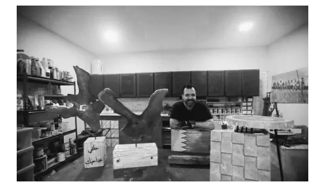 Foto van een glimlachende man in een werkplaats achter houten meubelstukken en een houten vogelsculptuur.