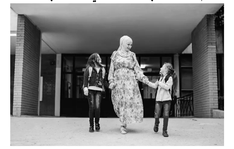 Foto van een vrouw met hoofddoek die hand in hand met twee meisjes een schoolgebouw uit loopt.