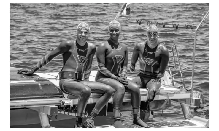 Foto van drie zwemmers in wetsuits die naast elkaar op de rand van een vlot op het water zitten.