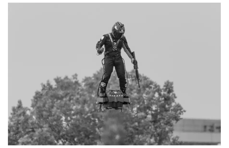 Foto van Franky Zapata die met een geweer in zijn hand op zijn Flyboard boven de bomen zweeft.