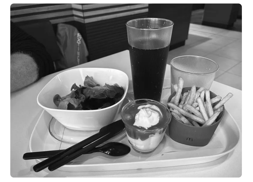 Foto van een fastfooddienblad met een kom salade, een glas drank, een bakje saus, frietjes en bestek.