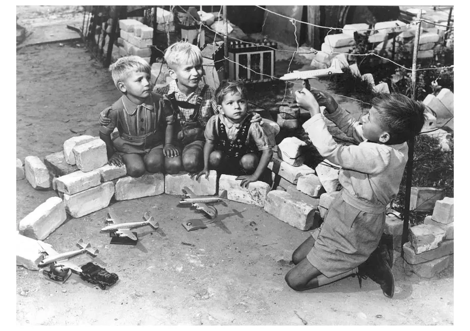 Foto van kinderen die met speelgoedvliegtuigjes en stenen de luchtbrug naspelen.