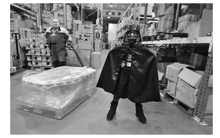 Foto van een magazijn met een verpakte pallet, en een jongen in een zwart Darth Vader-kostuum met cape en helm.