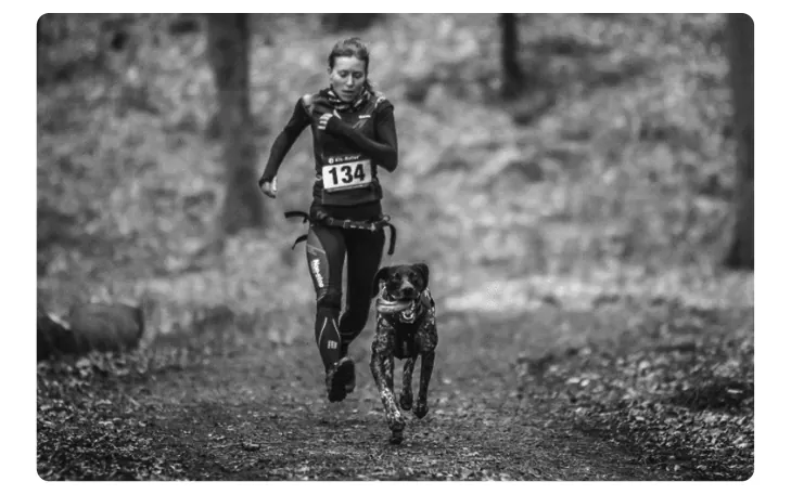 Foto van een hardloopster met startnummer 134 die samen met een hond aan een lijn over een bospad rent.