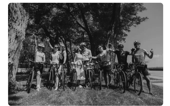 Groepsfoto van het bruidspaar en gasten met fietsen en helmen onder de bomen, sommigen zwaaien.