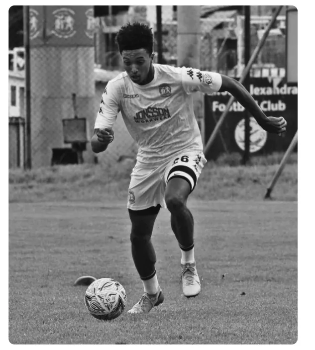 Foto van een jonge voetballer in een wit tenue die met de bal aan de voet over een grasveld loopt.
