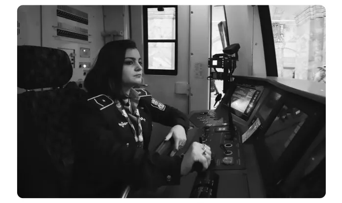 Foto van een vrouwelijke metrobestuurder in uniform in de cabine van een metrotrein, met haar hand op de bedieningshendel.