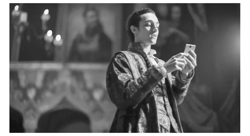 Foto van een man in een historisch kostuum die lachend naar zijn telefoon kijkt, op een toneel met een achtergrond van andere acteurs.