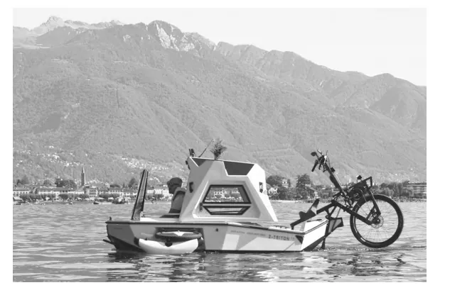 Foto van een fiets-amfibie op het water, met een lange bergketen op de achtergrond; de boot heeft een fiets aan de achterkant.