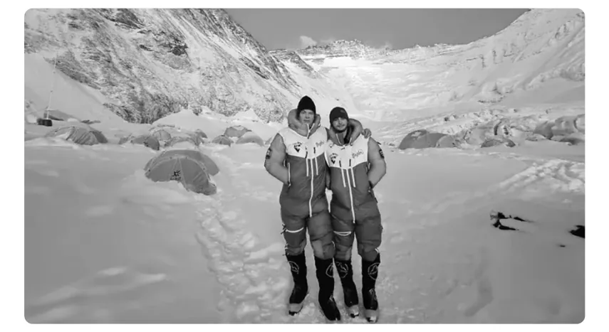 Zwart-witfoto van twee klimmers in dikke pakken op een besneeuwde vlakte, met tenten op de achtergrond.