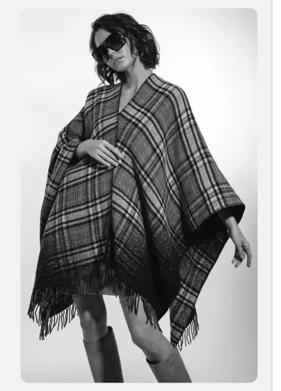 Foto van een vrouw met een geruit poncho met franjes, staand tegen een lichte achtergrond.