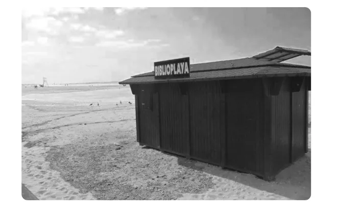 Zwart-witfoto van een donker houten strandhuisje met het bord BIBLIOPLAYA op een zandstrand.