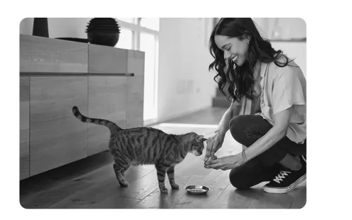 Zwart-witfoto van een vrouw die in een woonkamer op de vloer hurkt en een kat eten geeft.