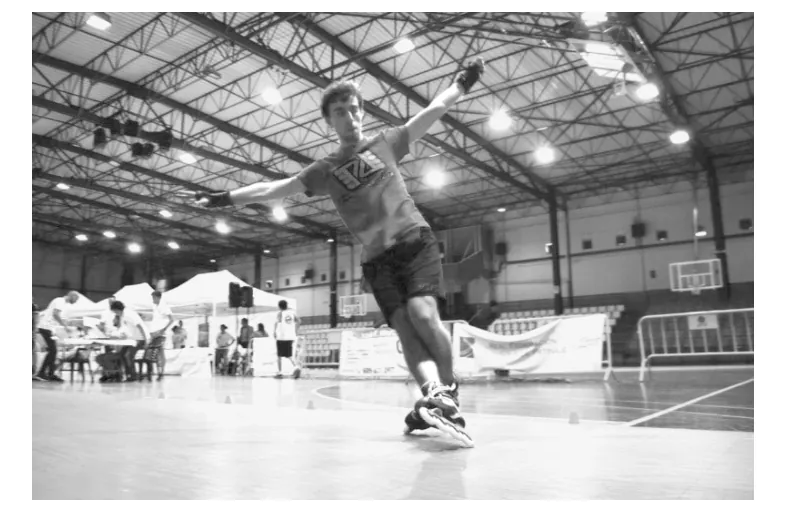 Foto van een man op inline skates die in een sporthal een sprong maakt, met op de achtergrond tribunes en tafels.