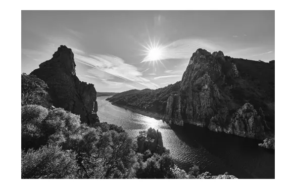 Zwart-witfoto van een rivier tussen rotsachtige heuvels met struiken, met de zon laag aan de hemel.