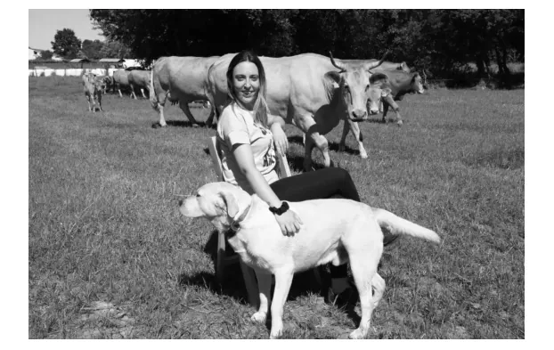 Zwart-witfoto van een jonge vrouw op een stoel in een weiland met een hond naast zich en koeien op de achtergrond.