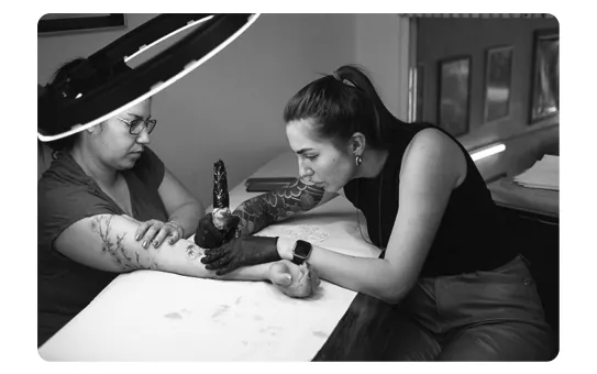 Zwart-witfoto van een tatoeëerder die aan de arm van een klant werkt.