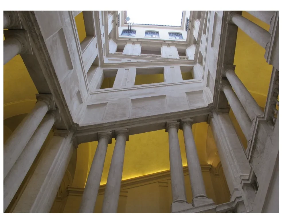 Blik omhoog in een trappenhuis met zuilen en lichte wanden, de trap in het midden en daarboven de ramen van de verdiepingen.