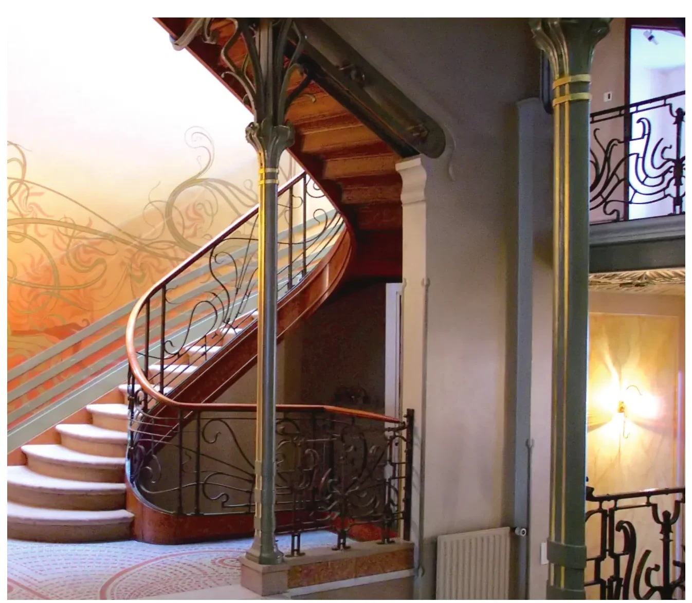 Trap met een smeedijzeren balustrade met krullende motieven, een houten leuning en een oranje geschilderde wand met patronen.