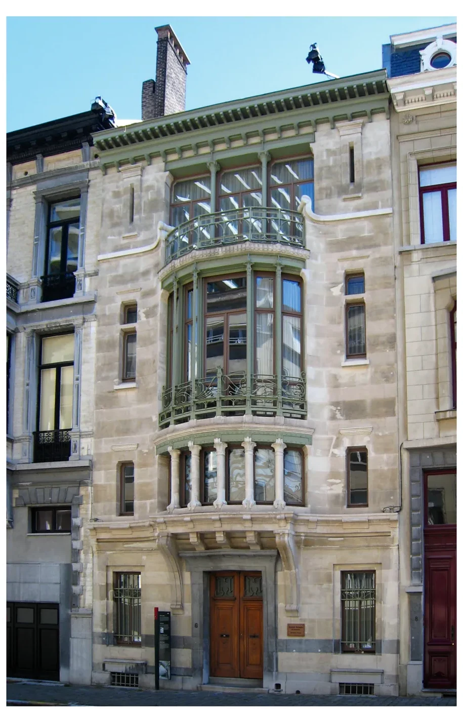 Voorgevel van Huis Tassel: een natuurstenen herenhuis met een erker met glas-in-lood en ijzeren ornamenten, een rij vensters en een donkere voordeur.