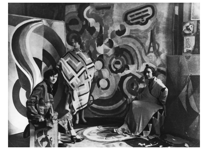 Zwart-witfoto in het atelier van Robert Delaunay: drie vrouwen voor een groot schilderij met cirkels, van wie een vrouw staand een geblokte mantel draagt.