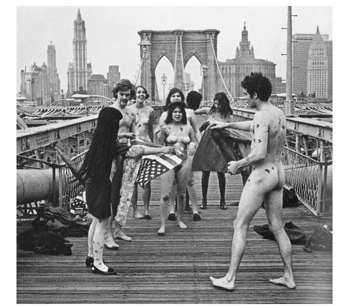 Zwart-witfoto van een happening op een brug in New York: naakte deelnemers tussen voorbijgangers, een vrouw links met een Amerikaanse vlag en de skyline op de achtergrond.