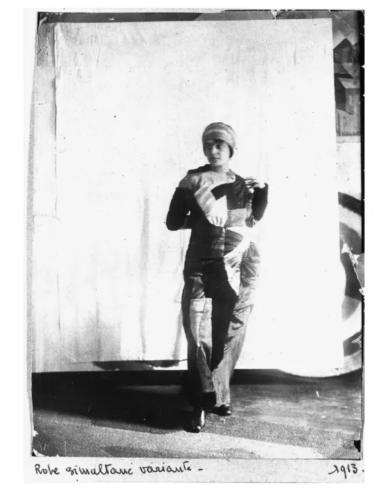 Zwart-witfoto van Sonia Delaunay in haar zelfgemaakte jurk, staand voor een lichte achtergrond, met een hoofddeksel en een jurk van verschillende stukken stof.