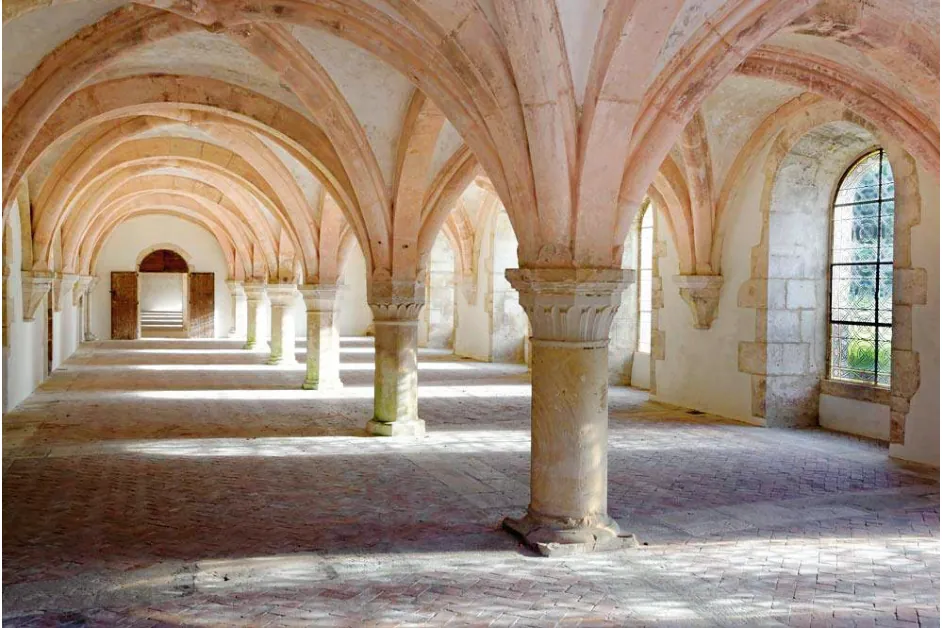 Lichte zaal met gewelven die rusten op korte, dikke zuilen, een stenen vloer en zonlicht dat door de ramen rechts naar binnen valt.