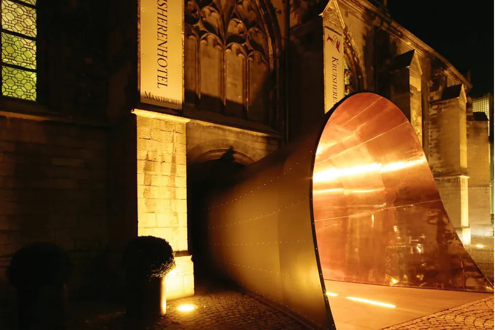 Avondopname van de koperen entreetunnel die schuin uit de gotische zijmuur van de kerk steekt; het glanzende koper licht warm oranje op.