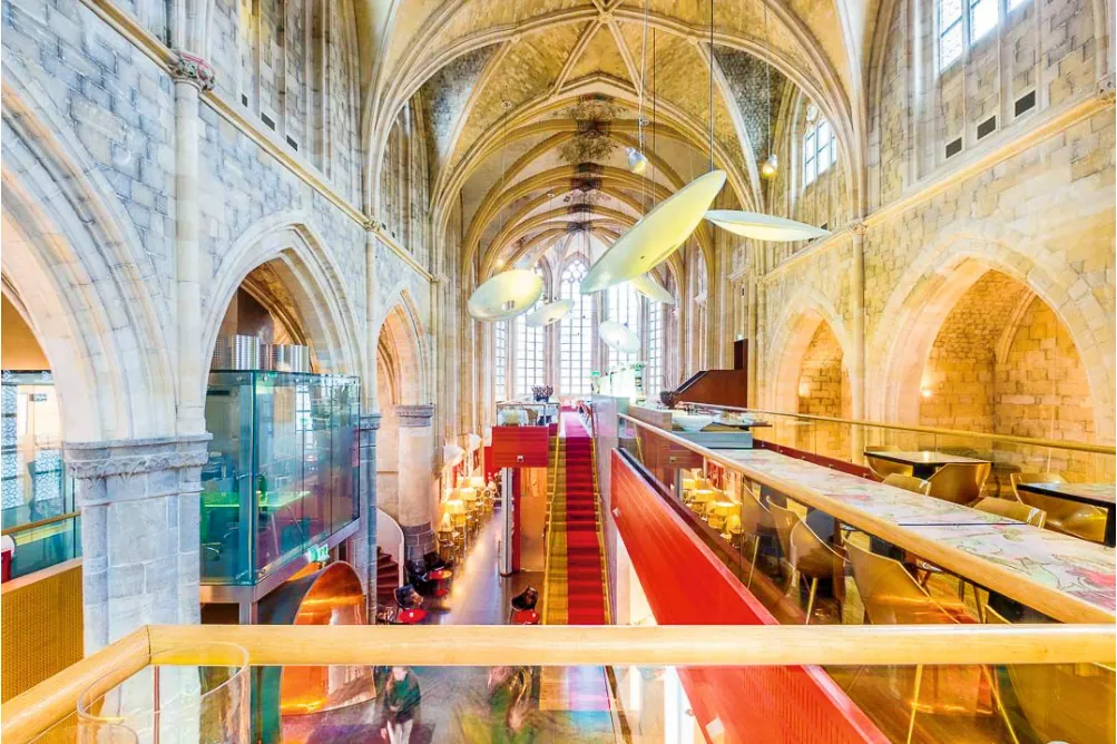 Het gotische schip van de kerk met een moderne tussenverdieping waarop het restaurant staat, een rode trap, een glazen lift en grote witte hanglampen onder de gewelven.