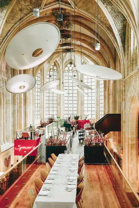 Het restaurant op de tussenverdieping met een lange gedekte tafel, met boven de tafels grote schotelvormige lampen en op de achtergrond het hoge gotische koorraam.