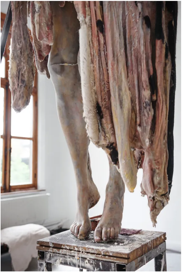 Detail van de benen van een figuur die op de tenen op een kleine sokkel staat, met lange gerafelde huiden in roze, bruin en zwart die langs de benen afhangen.