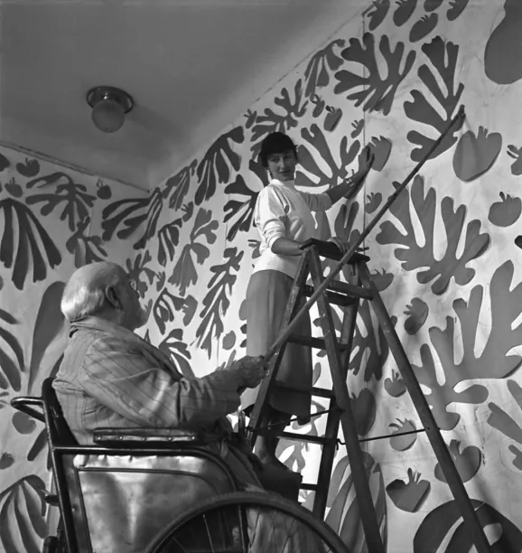 Zwart-witfoto van Matisse in een rolstoel in zijn atelier; een assistente op een trapje bevestigt met een lange stok knipsels van bladvormen op de wand, die al vol hangt met zulke vormen.