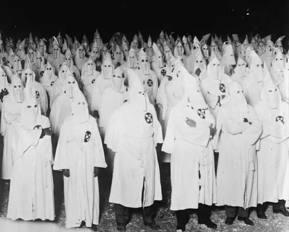 Zwart-witfoto van een grote groep mensen in witte gewaden met witte puntkappen die het gezicht bedekken, dicht op elkaar in de nacht.
