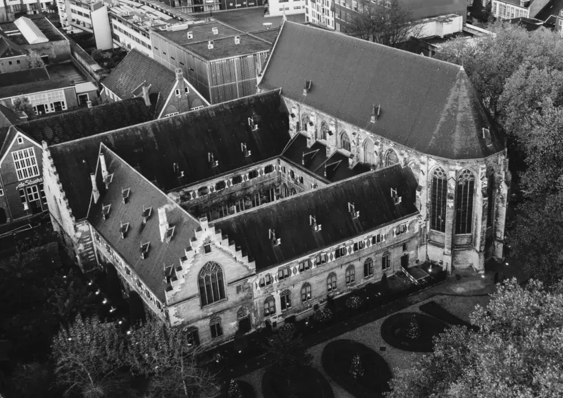Zwart-witte luchtfoto van het kloostercomplex van de Kruisheren: een hoge kerk met steil dak en grote ramen, en daaraan vast kloostervleugels met dakkapellen rond een binnenhof.