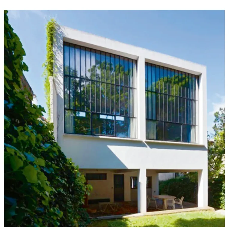 Achtergevel van de atelierwoning met grote raampartijen met roosters, een witte vlakke gevel en een balkon, omgeven door groen en blauwe lucht.