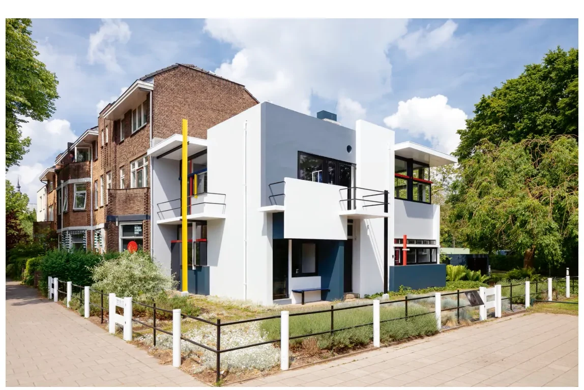 Het Rietveld-Schröderhuis vanaf de straat: witte gepleisterde muurvlakken, een plat dak met grote raampartijen en een gele lijn langs het balkon, naast een traditioneel bakstenen woonhuis.